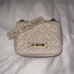 Beige quiltad axelväska från Love Moschino - Säljer en snygg beige axelväska från Love Moschino med quiltat mönster och guldfärgade detaljer. Väskan har en kedjerem och en praktisk dragkedja inuti. Perfekt för att ge din outfit en lyxig touch.
