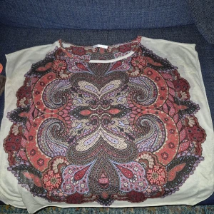 Mönstrad blus från Desigual - Säljer en färgglad blus från Desigual med ett detaljerat paisleymönster i rött, lila och beige. Blusen har en rund hals och är kortärmad, perfekt för att liva upp garderoben. Passar bra till både jeans och kjol!