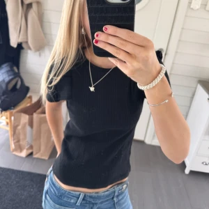 Svart ribbad topp med volangdetaljer❣️ - Snygg svart ribbad topp med volangdetaljer på axlarna. Toppen har en tight passform och korta ärmar, perfekt för en stilren look.