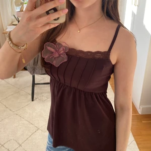 Brun vintage topp med spetsdetaljer - Säljer en söt brun topp med smala axelband och spetskant🤩Toppen har en rosa blomma på bröstet och en lätt rynkad midja🥰hör av er vid funderingar!!💕💕