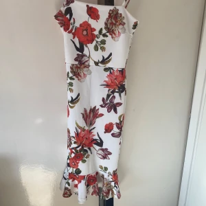 Vit blommig klänning - Säljer en vacker vit klänning med stora blommönster i rött och grönt. Klänningen har smala axelband och en volangdetalj vid fållen. Perfekt för sommarens festligheter!