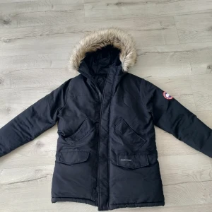 Svart parkajacka från Canada Goose - Säljer en svart parkajacka från Canada Goose med pälsfodrad huva. Jackan har en broderad logotyp på ärmen och praktiska fickor framtill.