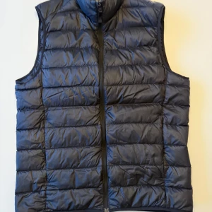 Svart dunväst från Uniqlo - Säljer en stilren och lätt svart dunväst från Uniqlo. Västen har en dragkedja framtill och är perfekt för lager-på-lager under kyliga dagar. Den är designad för att vara ultralätt och bekväm att bära.