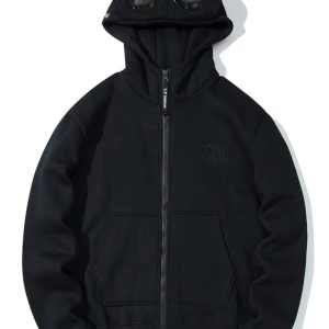 Svart hoodie med glasögon i huvan från C.P. Company - Säljer en svart hoodie från C.P. Company med unika glasögon i huvan. Den har en dragkedja framtill och en broderad logga på bröstet. Perfekt för en cool och avslappnad stil.