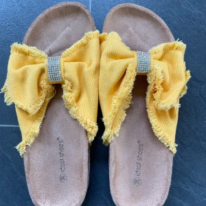 Gula sandaler med rosett och strassdetalj - Snygga gula sandaler  med en stor rosett och glittrande strassdetalj på ovansidan. Sandalerna har en bekväm sula och en fransig kant som ger en trendig look. Perfekta för sommarens alla äventyr!