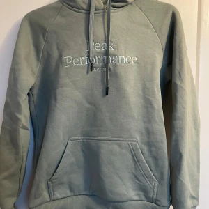 Grön hoodie från Peak Performance - Säljer en stilren grön hoodie från Peak Performance med broderad logga på bröstet. Tröjan har en klassisk känguruficka och justerbar huva med dragsko. Perfekt för en avslappnad look.