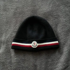 Svart mössa från Moncler - Snygg svart mössa från Moncler med ränder i vitt och rött. Den har en broderad logotyp framtill och är perfekt för kyliga dagar. Äkta, kommer inte till användning. 