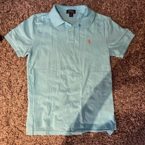 Ljusblå pikétröja från Ralph Lauren - Säljer en stilren ljusblå pikétröja från Ralph Lauren med klassisk krage och korta ärmar. Tröjan har en broderad logotyp i orange på bröstet. Perfekt för en avslappnad och snygg look. Nypris 1500kr