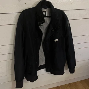 Svart jacka från G-Star RAW - Snygg svart jacka från G-Star RAW med hög krage och dragkedja. Jackan har en cool patch på ärmen och en knappdetalj på bröstfickan. Perfekt nu till våren🌸 En liten fläck inne i jackan men inget som syns ut.