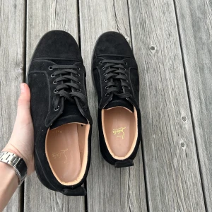 Christian Louboutin Svarta Sneakers - Snygga svarta sneakers i mocka från Christian Louboutin med klassisk röd sula. Skorna har snörning och en stilren design som passar perfekt för en trendig look.