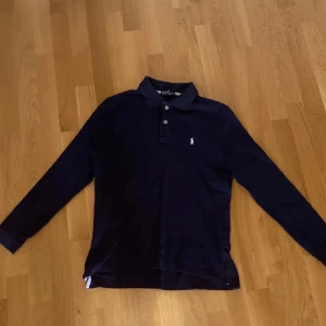 Blå Ralph Lauren pikétröja - Säljer en stilren svart långärmad pikétröja från Ralph Lauren med klassisk krage och knappdetaljer. Perfekt för en avslappnad men ändå uppklädd look. Bra skick, inga defekter.