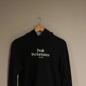 Svart hoodie från Peak Performance - Säljer en stilren svart hoodie från Peak Performance med vit text på framsidan. Perfekt för en avslappnad look. Den har en klassisk passform och en bekväm huva.