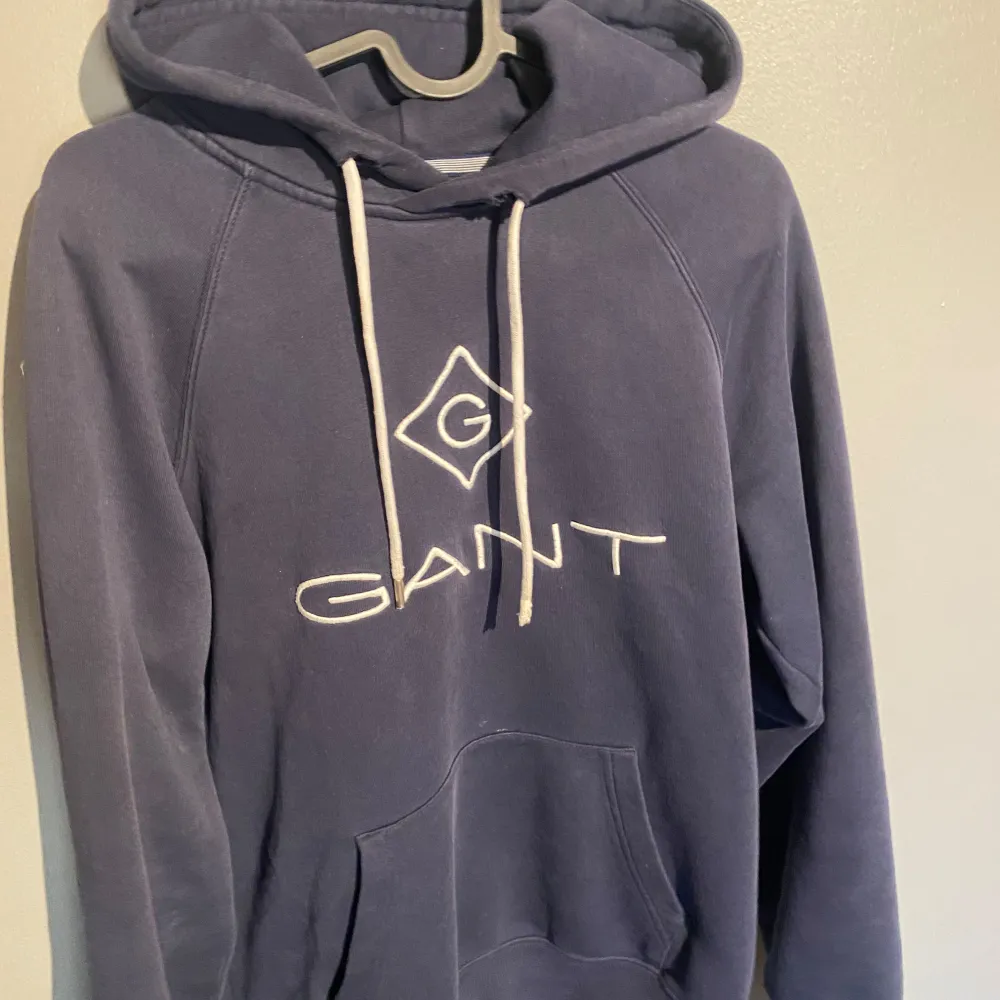 Säljer en snygg blå hoodie från GANT med vit logga på bröstet. Tröjan har en klassisk känguruficka och justerbar huva med vita snören. Perfekt för en avslappnad stil.. Neuletakit & Villapaidat.
