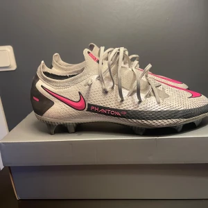 Nike Phantom GT fotbollsskor - Säljer ett par Nike Phantom GT fotbollsskor i grått med rosa detaljer. Skorna har snörning och en modern design med Nike-loggan på sidan. Perfekta för fotbollsplanen med sin avancerade greppteknologi.