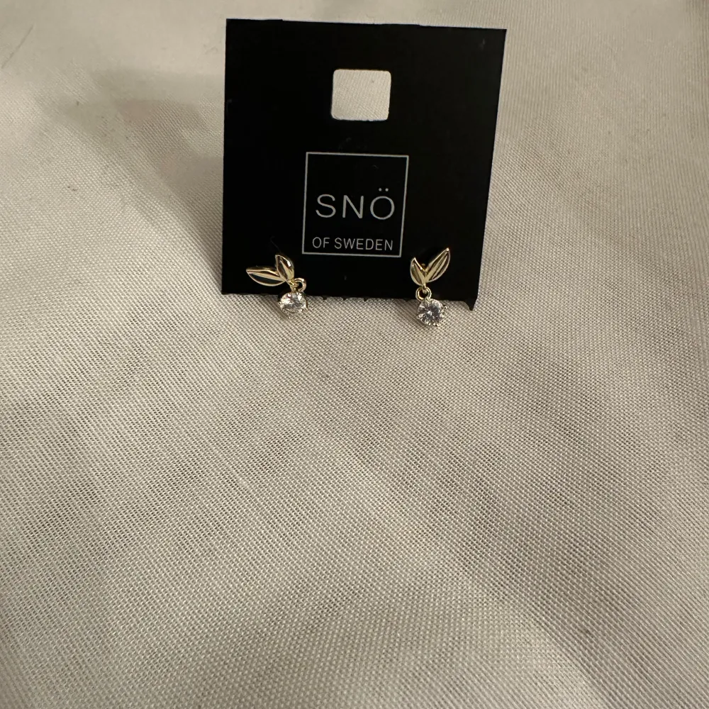 Eleganta örhängen från SNÖ of Sweden med en klar sten och guldfärgad detalj i form av ett löv. Perfekta för att ge en subtil glans till din look.. Asusteet.