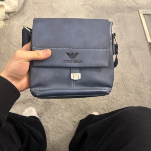 Blå axelväska från Giorgio Armani - Snygg blå axelväska från Giorgio Armani med magnetknappar och justerbar axelrem. Väskan har en stilren design med logotyp på framsidan och en praktisk ficka under locket. Perfekt för att bära dina nödvändigheter med stil. Pris går och diskutera 