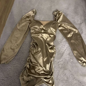 Champagnefärgad satin klänning - Snygg champagnefärgad klänning i satin med långa puffärmar och en v-ringad design. Klänningen har en tight passform och är kort, perfekt för en festlig kväll. Den glänsande ytan ger en lyxig känsla.