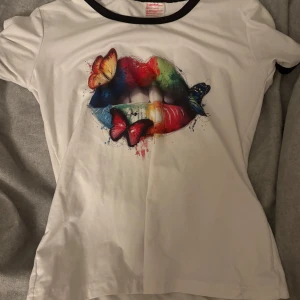Vit t-shirt med färgglatt läpp tryck - Säljer en vit t-shirt med ett coolt färgglatt tryck av läppar och fjärilar. T-shirten har svarta kanter vid halsen och är kortärmad. Perfekt för en färgstark look! 🌈🦋