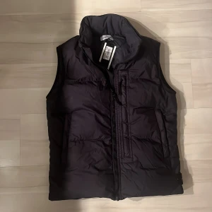 Marinblå dunväst från Stone Island - Snygg marinblå dunväst från Stone Island med dragkedja och broderad logga på sidan. Luva finns men kan vikas in. Perfekt för att hålla värmen under kyliga dagar. Västen har en stilren design och är lätt att matcha med olika outfits. Storlek 170/14 år. Helt oanvänd lappar kvar!