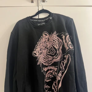 Svart tröja med tigertryck från Philipp Plein - Säljer en svart tröja från Philipp Plein med ett stort tigertryck i rosa på framsidan. Tröjan har rund hals och ett metallmärke med PP-logga på baksidan. Perfekt för dig som vill ha en statement piece i garderoben!