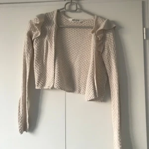 Beige kofta från H&M - Säljer en söt beige kofta från H&M med volangdetaljer vid axlarna. Koftan är stickad med ett fint mönster och har långa ärmar. Perfekt för lager-på-lager under kyligare dagar. 🧥✨