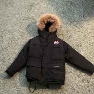 Svart dunjacka med pälsdetalj - Säljer på grund av att den är för liten för mig. Säljer en svart dunjacka från Canada Goose med avtagbar pälsdetalj på huvan. Jackan har flera praktiska fickor och en dragkedja framtill. Perfekt för kalla vinterdagar.Den passar nog en 10-12 åring.Längd 57cm, armar 47cm