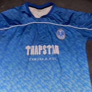 Blå fotbollströja från Trapstar - Säljer en blå fotbollströja från Trapstar med ett unikt mönster och vit text. Tröjan har korta ärmar och nummer 22 på ryggen. Perfekt för dig som vill sticka ut på planen eller bara gillar stilen. 🏆