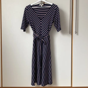 Randig klänning från Hampton Republic - Vackert figursydd randig klänning från Hampton Republic i marinblått, vitt och rött. Klänningen har en v-ringad hals och trekvartsärmar. Den är knälång och har ett knytband i midjan för en fin passform och mycket vidd i kjolen. Mjukt material med mycket stretch. Längd 110, mått runt midjan 84. 