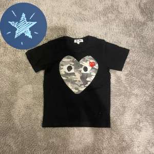 Cdg T-shirt  - Snygg svart t-shirt från PLAY med ett stort camo-mönstrat hjärta på bröstet. Perfekt för en cool och avslappnad stil.