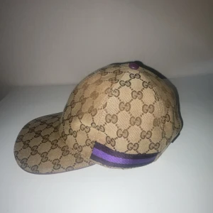 Beige keps med GG-mönster - Snygg beige keps från Gucci med det ikoniska GG-mönstret. Kepsen har en justerbar rem baktill och en lila detalj på sidan. Perfekt för att ge din outfit en lyxig touch.