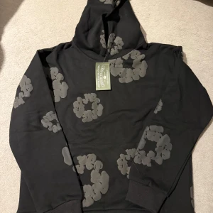 Svart hoodie från Denim Tears - Säljer en svart hoodie från Denim Tears med unikt blommönster i grått. Hoodien har en klassisk passform med långa ärmar och ribbade muddar. Perfekt för en stilren och bekväm look.