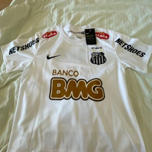 Nike Santos tröja Neymar 11 - Säljer en vit fotbollströja från Nike med Santos FC-logga och sponsortryck i svart och guld. Tröjan har korta ärmar och spelarens namn och nummer på ryggen. Perfekt för fotbollsfans!