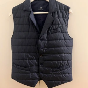 Mörkblå quiltad väst från Hansen jacob  - Snygg mörkblå quiltad väst från Hansen jacob med knappar framtill. Västen har en klassisk krage och flera fickor för praktisk användning. Perfekt för att hålla stilen när det är lite kyligare ute.