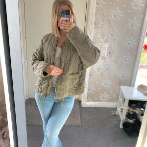 Olivgrön quiltad jacka - Säljer en olivgrön quiltad jacka med dragkedja och rund hals. Jackan har en avslappnad passform och är perfekt för vår och höst dagar. 