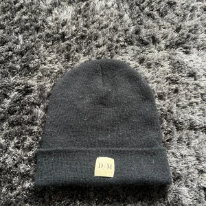 Svart mössa med etikett - Snygg svart mössa med uppvikt kant och en liten etikett med initialerna D/M framtill. Perfekt för kyliga dagar och enkel att matcha med olika outfits.