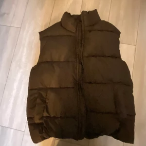 Svart dunväst från Monki - Säljer en brun dunväst från Monki. Västen har en dragkedja framtill och är perfekt för kyligare dagar. Den är stilren och lätt att matcha med olika outfits. Passar bra för både vardag och utomhusaktiviteter.