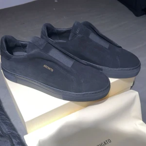 Svarta Arigatos - Snygga svarta sneakers från Axel Arigato i en stilren design. Skorna är tillverkade i mocka och har en diskret logga i guld på sidan. Perfekta för en minimalistisk look.