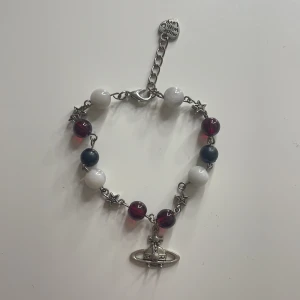 Snyggt stjärn armband med vivienne westwood detalj - Charmigt armband med vita, röda och blå pärlor samt små stjärndetaljer. Armbandet har en silverfärgad kedja och ett hänge med en unik design. Perfekt för att ge en färgklick till din outfit. ‼️Köpte den på plick inte egengjord‼️