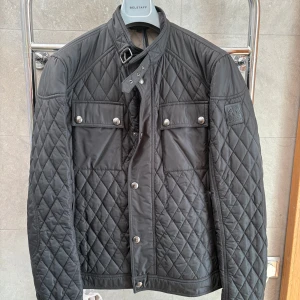 Snygg svart quiltad jacka från Belstaff - Snygg svart quiltad jacka från Belstaff med dragkedja och knappar. Jackan har en stilren design med en hög krage och två bröstfickor med knappar. Perfekt för en cool och avslappnad look.