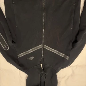 Svart hoodie och byxor från Nike tech  - Säljer en svart hoodie plus byxor från  Nike med dragkedja och huva. Den har en diskret logga på bröstet och ärmarna. Perfekt för en sportig look eller avslappnade dagar. Passar 147-158 cm 