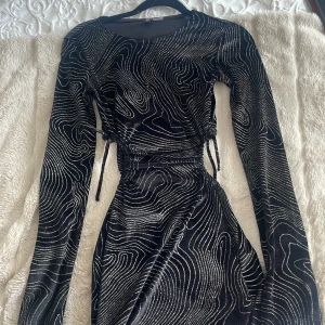 Svart klänning från H&M Divided - Snygg svart klänning från H&M Divided med ett unikt mönster i silver. Klänningen har långa ärmar och en åtsittande passform med knytning i midjan. Perfekt för en kväll ute eller en speciell tillställning.