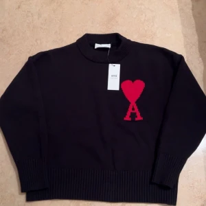Svart stickad tröja från Ami - Säljer en snygg svart stickad tröja från Ami med ett rött hjärta och bokstaven A på bröstet. Tröjan har en ribbad krage och långa ärmar. Perfekt för att hålla sig varm och stilren.