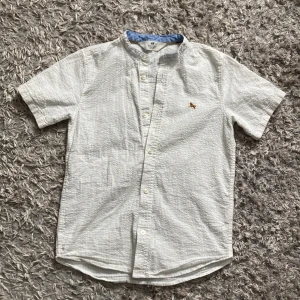 Vit kortärmad skjorta t-shirts med knappar  - Säljer en vit T-shirt/skjorta linne storlek 134 med ett märke på bröstet och med knappar 