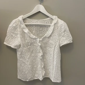 Vit blus med volangdetaljer - Charmig vit blus med korta ärmar och volangdetaljer längs kragen och framsidan. Blusen har ett vackert broderat mönster som ger en romantisk känsla. Perfekt för en somrig look!