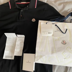 Svart pikétröja från Moncler FINNS KVAR 08/01 - Säljer en stilren svart pikétröja från Moncler med klassiska röd-vita detaljer på krage och ärmslut. Tröjan har en broderad Moncler-logga på bröstet och knappar framtill. Perfekt för en avslappnad men ändå elegant look. Den är storlek L men passar som M. Den är köpt 2022 i milano, men är knappt använd och det finns två små hål på framsidan som du kan se på sista bilden. Kvitto, original paket och allt annat finns med, finns även veriferings kod.