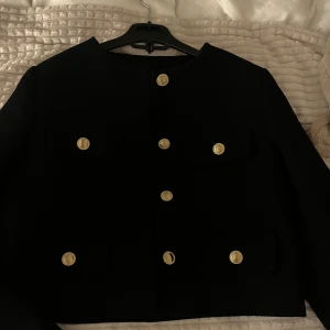Svart vår/sommar jacka från Zara - Snygg svart kavaj från Zara med guldknappar. Den har en klassisk design med rund hals och långa ärmar. Perfekt för en stilren look.