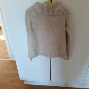 Beige stickad tröja med polokrage - Mysig beige stickad tröja med en bred polokrage. Perfekt för kyliga dagar med sin varma och bekväma design. Tröjan har långa ärmar och ribbade detaljer vid krage, ärmslut och nederkant.