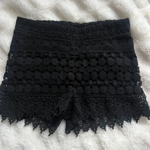 Svarta spets shorts - Snygga svarta shorts i spets med detaljerade mönster. Perfekta för en stilren look med en touch av elegans. De har en högmidjad passform och en dragkedja bak för enkel påklädning.