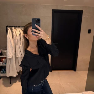 Svart blus med volangdetaljer - Snygg svart blus med volangdetaljer och knappar framtill. Perfekt för en stilren look. Blusen har långa ärmar och en elegant design som passar till många tillfällen.