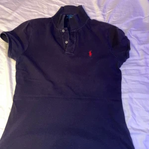 Mörkblå pikétröja från Ralph Lauren - Säljer en klassisk mörkblå pikétröja från Ralph Lauren med korta ärmar och en röd broderad logotyp på bröstet. Perfekt för en stilren och avslappnad look. Tröjan har en knappslå med två knappar vid halsen. Den är i storlek s och passar väldigt bra på